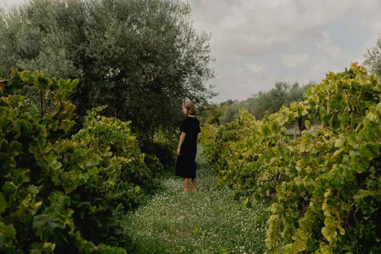 Vrouw in zwarte jurk wandelt tussen groene wijngaarden onder bewolkte lucht, natuurlijke sfeer voor huidverzorging